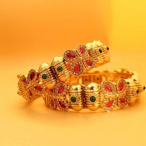 Può includere: Due braccialetti d'oro con disegni intricati che presentano pietre preziose rosse e verdi. I braccialetti sono impilati uno sopra l'altro e hanno un motivo floreale.