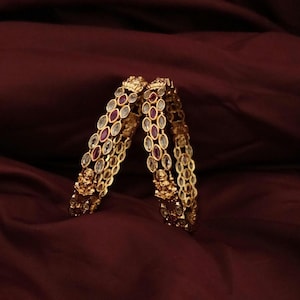 May include: Two gold-toned bangles featuring a repeating marquise-shaped pattern set with clear white stones and alternating deep red stones. Each bangle includes a prominent gold-toned figure of a deity integrated into the metalwork design.