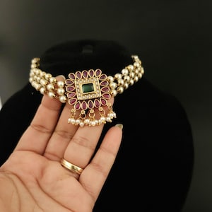 Könnte beinhalten: Ein goldfarbenes Choker-Halsband mit drei Strängen aus kleinen, weißen Perlen. Der Mittelteil zeigt einen quadratischen grünen Stein, rote Edelsteine und goldene Details, mit kleinen Perltropfen. Die Halskette wird auf einer schwarzen Schaufensterpuppe präsentiert.