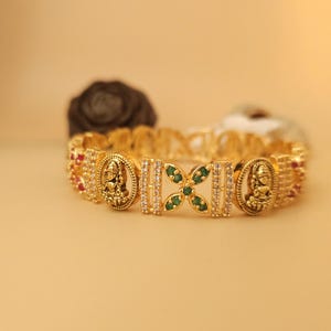 Op de afbeelding: Een gouden armband met een decoratief ontwerp met kleine witte stenen, groene stenen en twee gouden figuren.