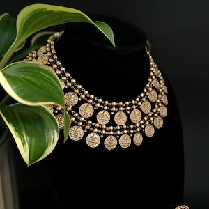 May include: A multi-layered gold-toned necklace featuring three rows of small, round, embossed coin-like charms. The necklace is displayed on a black bust, with each chain connected by small, polished gold-toned beads between the circular pendants.