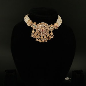 Puede incluir: Un collar dorado adornado con un colgante central circular con detalles intrincados y piedras preciosas de colores. El colgante cuelga de un cordón de cuentas blancas. El collar se exhibe en un maniquí negro.
