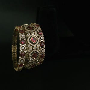 Peut inclure: Deux bracelets dorés avec des détails complexes. Chaque bracelet est orné de nombreux pierres rouges et de petites pierres transparentes, selon un motif répétitif. Les bijoux sont présentés sur un fond sombre.