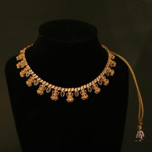 May include: A gold-toned necklace featuring a series of repeating deity figures and oval-shaped dark green stones. The chain is composed of small, linked metallic segments, and the piece includes a thin, adjustable cord with a small beaded tassel.