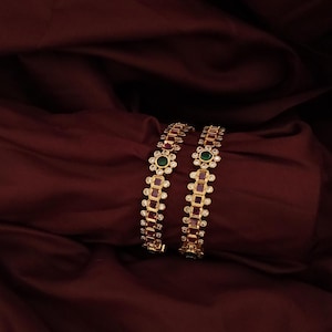 May include: Two gold-tone bangles featuring a repeating pattern of rectangular deep red stones and circular clear stones. Each bangle includes a central flower-shaped motif with a round green stone at the centre, surrounded by small clear stones.
