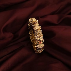 May include: A gold-toned bangle bracelet featuring intricate embossed relief carvings of a seated deity figure. The metal surface is decorated with small, embedded red and green stones and clear pavé-style accents along the circular band.