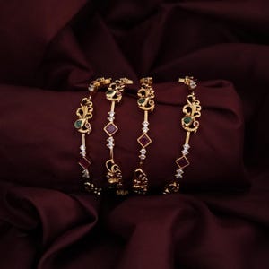 May include: A set of four gold-tone bangles featuring intricate metalwork, small clear stones, square-cut deep red stones, and teardrop-shaped green stones. The bangles are arranged in a row, showcasing a repeating pattern of ornate metal motifs.