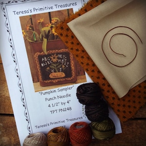 Może przedstawiać: Zestaw rzemieślniczy z wzorem igły do dziurkowania "Pumpkin Sampler". Zestaw zawiera drukowany wzór, tkaninę, nić i igłę. Wzór ma wymiary 11,4 cm na 10,2 cm. Kolory to brązowy, pomarańczowy i beżowy.