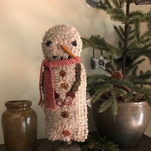 Könnte beinhalten: Ein gehäkelter Schneemann mit einem rot-weißen Schal, einer braunen Zweignase und braunen Knopfaugen. Der Schneemann steht auf einer Metallglocke.