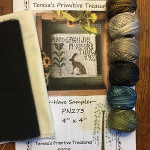 May include: A craft kit with a sampler design featuring a rabbit, flowers, and the alphabet. The kit includes a piece of black and white fabric, along with several spools of embroidery thread in various colors. The sampler is 4" x 4".