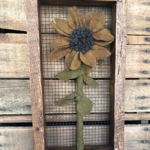 May include: A rustic wooden framed wall art piece featuring a large, textured sunflower. The sunflower has brown petals, a dark gray center, and green leaves and stem. The frame has the text "TERESA'S PRIMITIVE TREASURES" at the top.