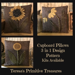 Puede incluir: Tres diseños diferentes para una almohada con un girasol, una margarita y una flor estilizada. Las almohadas están hechas de una tela marrón con un patrón sutil. El texto "Cupboard Pillows 3 in 1 Design Pattern Kits Available Teresa's Primitive Treasures" se muestra debajo de las almohadas.