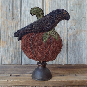 May include: A handcrafted Halloween decoration. It features a textured, rust-coloured pumpkin with green accents, topped by a black crow. The pumpkin is mounted on a dark metal base, creating a rustic, seasonal display.