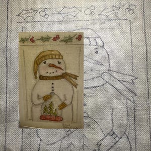 May include: A hand-painted illustration of a snowman holding a small Christmas tree in a snow globe. The snowman is wearing a brown hat and scarf. The illustration is surrounded by a border of holly with red berries.