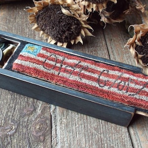 May include: A wooden box with a hinged lid, containing a needlepoint piece depicting an American flag with the words "God Bless" stitched in black thread. The box is painted black and has a distressed finish. The box is open and the needlepoint piece is visible. There are buttons and other sewing supplies in the box.
