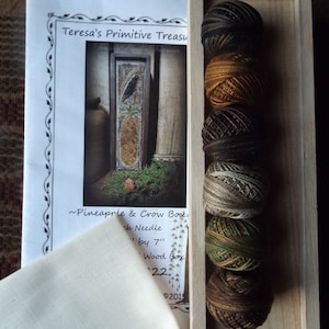 May include: A craft kit featuring a printed image of a pineapple and crow design, a piece of fabric, and several spools of thread in various earth-tone colours. The kit includes a wood box and a needle.