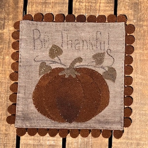 May include: A square, tan fabric decoration with a brown pumpkin and the words "Be Thankful!" stitched on it. The pumpkin has a stem and leaves. The border is trimmed with brown pom-poms. The decoration is likely for the fall season.