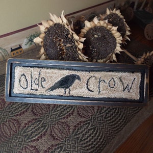 May include: A rectangular, framed sign with the words "Olde Crow" and a black crow illustration. The sign is made of a textured, beige material with a black border. Dried sunflowers are in the background.