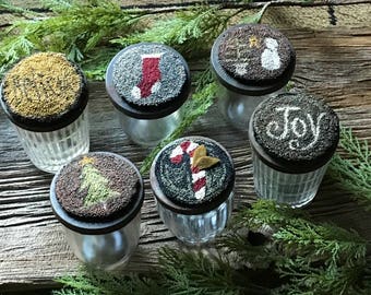 May include: Six clear glass jars with dark lids, each topped with a unique, textured design. The designs include a Christmas tree, a stocking, a snowman, a candy cane, a gold star, and the word "Joy".