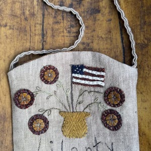 Könnte beinhalten: Schultertasche aus beigem Stoff mit einem aufgestickten Design mit einer amerikanischen Flagge, Blumen und dem Wort "Liberty". Die Tasche hat einen gewebten Griff und ist mit gestickten Details in Braun-, Gelb- und Blautönen verziert.