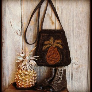 Puede incluir: Un bolso de hombro marrón con un diseño de piña, una piña fresca y un par de botas de cuero marrón con detalles de botones se exhiben sobre un fondo de madera. El bolso presenta un fondo marrón oscuro con un diseño detallado de piña.