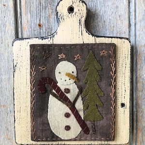 Peut inclure: Décoration murale sur le thème du bonhomme de neige. Le bonhomme de neige est en feutre blanc avec un nez en carotte et un sucre d'orge. Le fond est en tissu marron foncé avec une bordure cousue et un sapin de Noël en feutre vert. La planche en bois est peinte en crème.