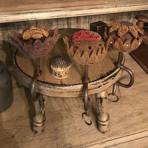 Puede incluir: Tres alfileteros de tela con forma de flores, cada uno con un diseño diferente, se muestran en un pequeño taburete de madera con tres patas. Los alfileteros son marrones con detalles dorados y rojos. El taburete está pintado de blanco con detalles negros.