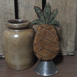 Puede incluir: Un adorno de punto de aguja en forma de piña de color marrón y verde con un patrón de cuadros marrones y blancos. El adorno se muestra en un soporte de metal plateado.
