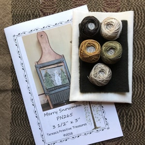 Op de afbeelding: Een naaldpuntset met een sneeuwpop met een groene boom en een houten frame. De set bevat vijf bollen borduurgaren in tinten bruin, beige, groen en goud. De set is getiteld "Merry Snowman PN265 3 1/2" x 3" Teresa's Primitive Treasures ©2018."