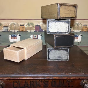 Puede incluir: Una pila de cuatro cajas de madera con cajones, pintadas en varios colores, incluyendo beige, blanco, negro y gris. Las cajas están apiladas sobre una mesa de madera con el texto "CLARK'S O.N.Z." impreso en la superficie.