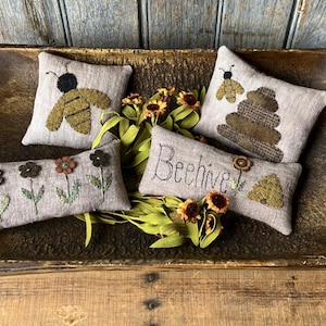 Può includere: Una ciotola di legno contiene quattro cuscini decorativi con ricami. Due cuscini presentano api, uno dice "Beehive", e un altro ha fiori ricamati. I cuscini sono in tonalità di grigio, marrone e verde.