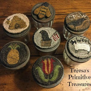 May include: A selection of glass jars with metal lids, each featuring a distinct, hand-stitched design. The designs include a bee, a pineapple, a tulip, a sheep, an eagle with a flag, a bird, and a bee. The jars are displayed on a wooden surface.