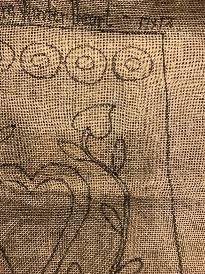 May include: A close-up of a burlap fabric with a black outline of a heart and a floral design. The fabric has the words "Winterheart" and "Mix" written on it.