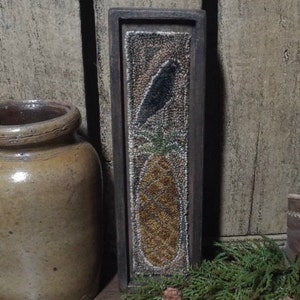 May include: A framed needlepoint artwork featuring a black bird perched on top of a pineapple. The pineapple is depicted in shades of brown, yellow, and green. The frame is dark brown wood.
