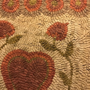 May include: A close-up of a handmade rug with a heart design in the center. The rug is made of small, colorful fabric scraps sewn together. The heart is surrounded by flowers and leaves.