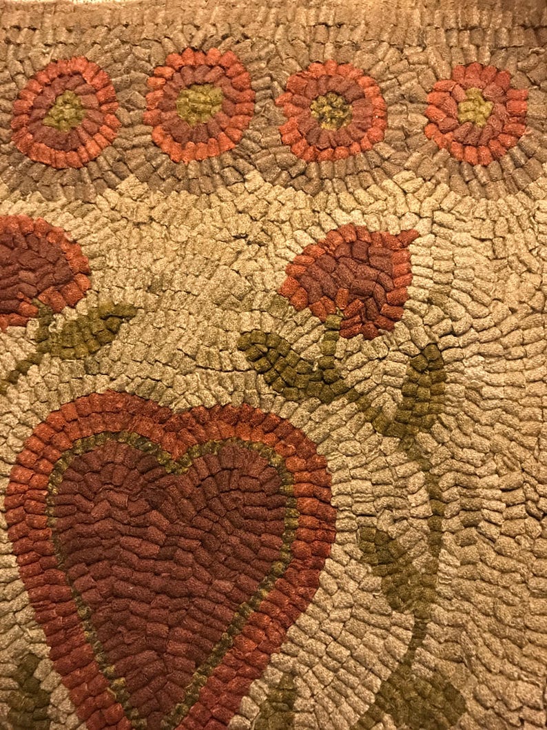 May include: A close-up of a handmade rug with a heart design in the center. The rug is made of small, colorful fabric scraps sewn together. The heart is surrounded by flowers and leaves.