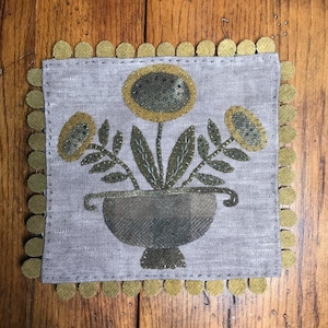 May include: A hand-stitched embroidery of three stylized flowers in a pot, with a brown and green color scheme. The flowers are surrounded by a gold felt trim.