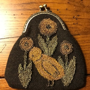 May include: A brown felt coin purse with a metal clasp. The purse is decorated with embroidered flowers and a bird in shades of yellow and green.