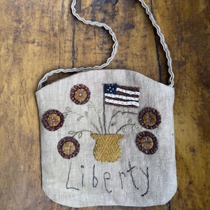 May include: Tan fabric bag with a stitched design featuring a small American flag, flowers, and the word "Liberty." The bag has a woven handle and decorative stitching around the edges. The flowers are brown and yellow.