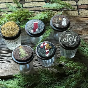 May include: Six glass jars with lids decorated with Christmas-themed needlepoint designs. The designs include a stocking, a candy cane, a Christmas tree, a snowman, a star, and the word "Joy".