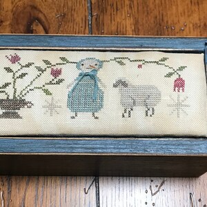 May include: A wooden box with a blue painted frame and a cream-colored fabric interior. The fabric features an embroidered design of a girl in a blue dress, a sheep, and a floral arrangement.