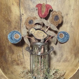 Pode incluir: Um pequeno pássaro amarelo com olhos castanhos senta-se num frasco de vidro com uma gaiola de metal. O frasco está cheio de musgo verde e o pássaro é rodeado por flores de feltro em tons de azul, vermelho e castanho.