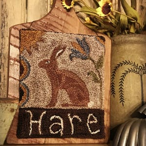 May include: A brown and tan needlepoint wall hanging with a brown rabbit and the word "Hare" in brown lettering. The hanging is framed in a wooden board with a handle.