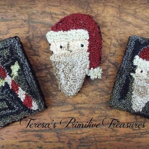 Può includere: Tre ornamenti natalizi in lana cuciti a mano su una superficie di legno. Gli ornamenti includono un volto di Babbo Natale e due disegni rettangolari, uno con un bastoncino di zucchero. Il testo "Teresa's Primitive Treasures" è visibile.