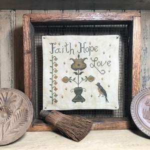May include: A framed cross-stitch sampler with the words "Faith Hope Love" and floral and bird motifs. The sampler is displayed in a rustic wooden frame with a wire mesh backing. Decorative wooden plates and a small broom are also present.