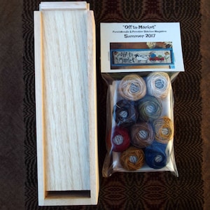 Puede incluir: Una caja de madera con una pequeña abertura en la parte delantera. Junto a la caja hay una bolsa de plástico que contiene un juego de hilo de bordar en varios colores. La bolsa tiene una etiqueta que dice "Off to Market" Punch Needle & Primitive Stitcher Magazine Summer 2017.
