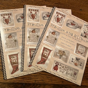 May include: Three spiral bound books with the title "Primitive & Folk Art Fancies" on the cover. The books contain designs in fiber for all seasons by Robin Leuschen and Teresa Miller. The cover of the book features a variety of primitive folk art designs, including a bird, a pumpkin, a stocking, a bee, and a Christmas tree.