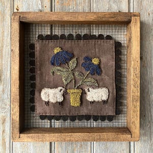 May include: A framed textile artwork. It depicts blue flowers, green leaves, and two sheep on a brown background. The flowers are in a yellow pot. The frame is made of wood.