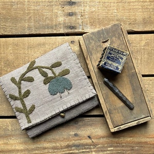 May include: A small, beige fabric pouch with a green floral design and a button closure. The pouch is sitting on a wooden surface next to a small wooden box with a blue and gold label that reads "Best Glass Head Pins The Royal Pin Co. 100 Pins".