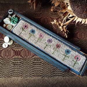 Puede incluir: Una caja de madera azul con un diseño floral sobre un fondo beige. La caja contiene varios botones y un marcapáginas bordado con flores. El fondo es una tela estampada con un girasol seco.